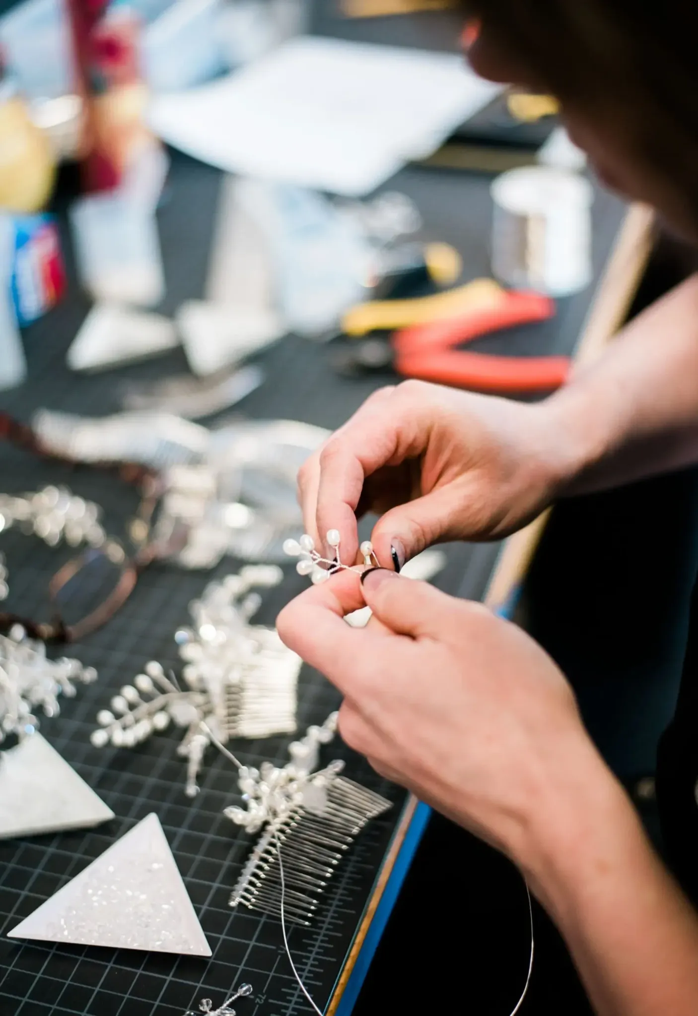 A person's hands crafting bead earrings on a busy workbench with jewelry making tools and materials