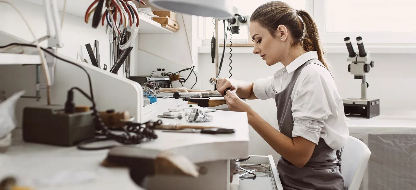 A woman working on jewelry at a cluttered workbench in a well lit room A woman working on jewelry at a cluttered workbench in a well lit room