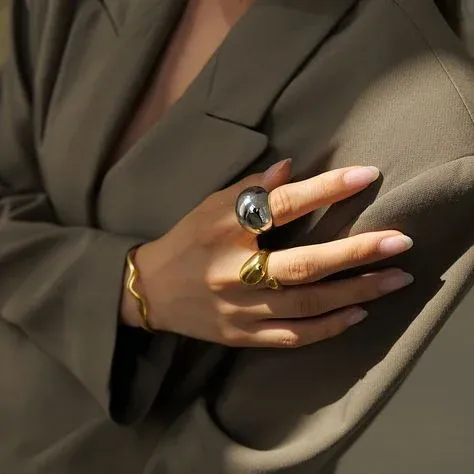 A person wearing a grey blazer, showcasing gold and silver rings and a gold bracelet A person wearing a grey blazer, showcasing gold and silver rings and a gold bracelet