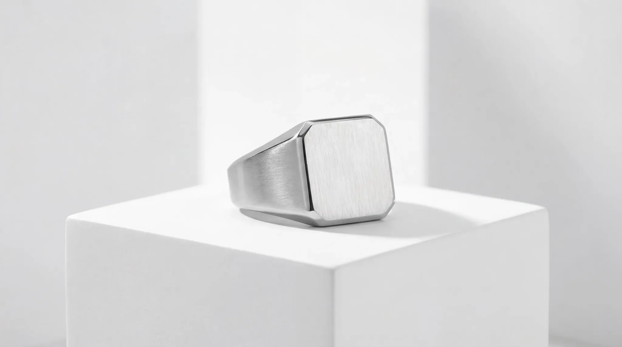 Minimalist square-faced stainless steel signet ring with a brushed matte finish on a white geometric pedestal