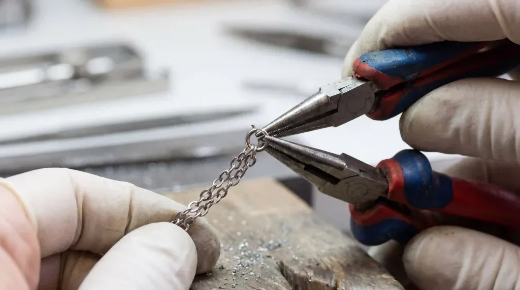 Close-up of needle-nose pliers being used to precisely close a small jump ring on a silver necklace chain.