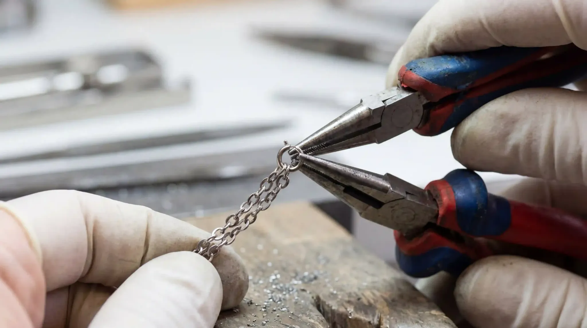 Close-up of needle-nose pliers being used to precisely close a small jump ring on a silver necklace chain.