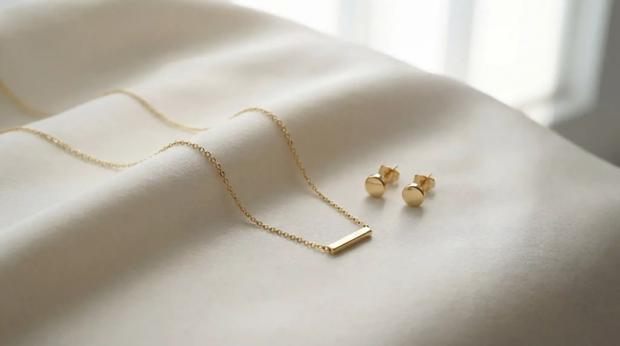 Minimalist gold necklace and stud earrings on a soft cream fabric