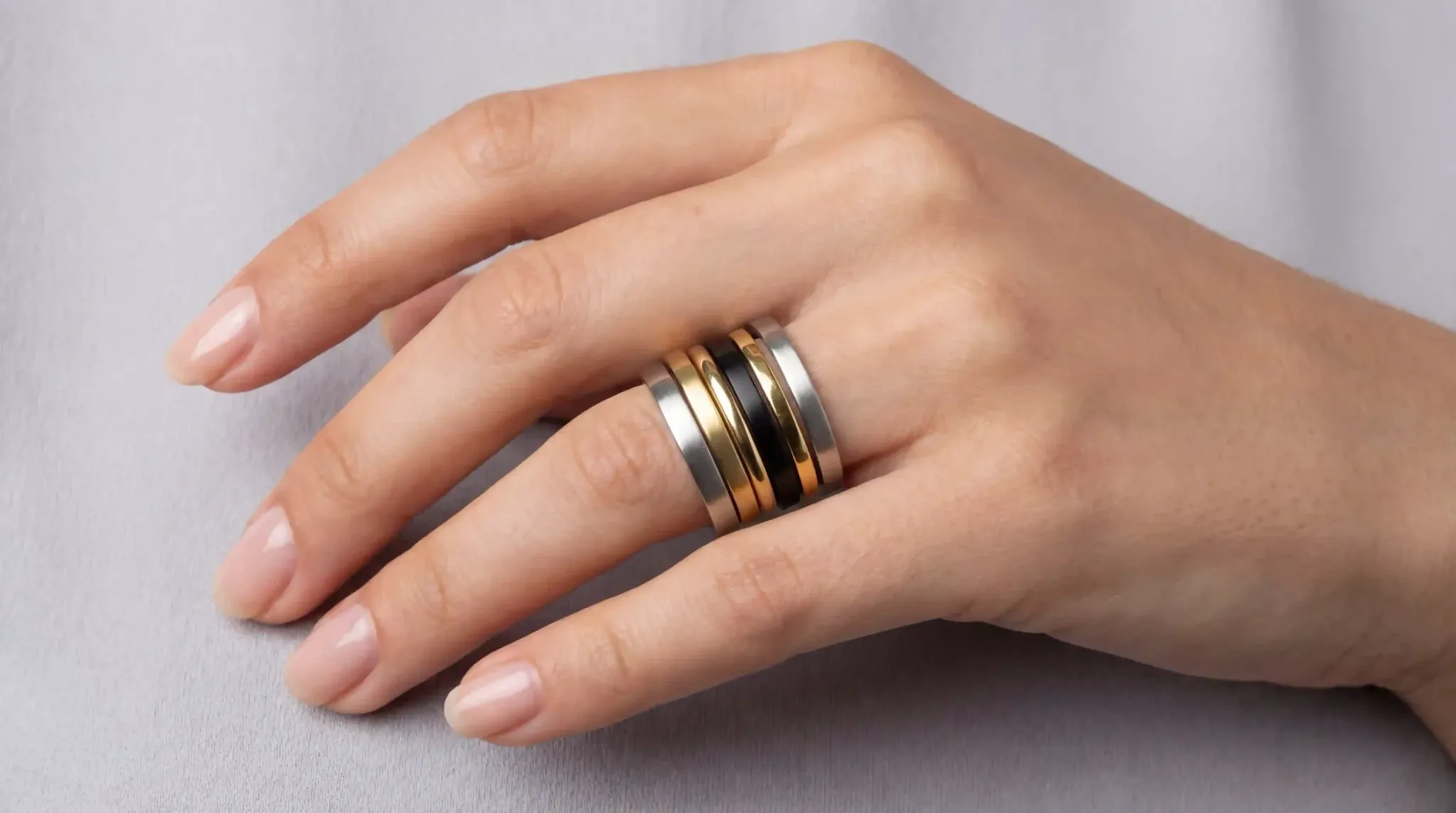 A hand wearing a stylish stack of five minimalist stainless steel bands in different finishes like gold and black.